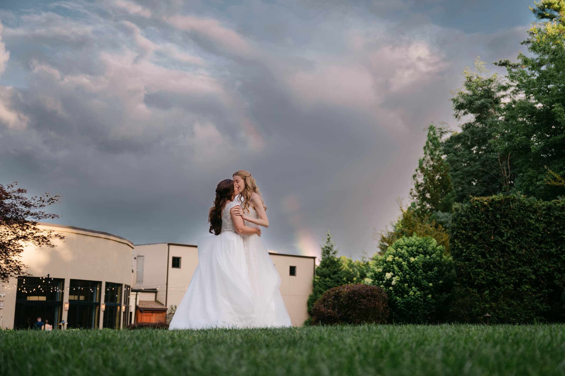 Two brides embracing on the lawn under a rainbow sky